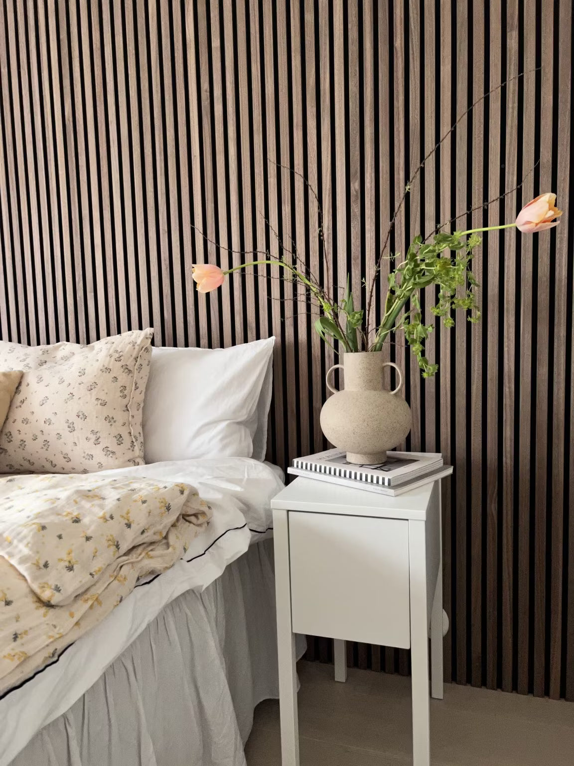 Une chambre à coucher avec un lit blanc, une literie florale et un vase de fleurs, avec un mur daccent en lattes acoustiques Akupanel en bois nordique derrière le lit.